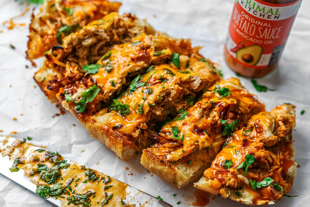 A loaf of buffalo chicken garlic bread sliced into 6 pieces next to a bottle of Primal Kitchen Buffalo Sauce. Knife and herbs in background. 