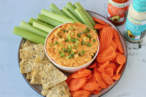 bowl of Buffalo Chicken Dip on a plate of crackers and veggies, next to bottles of Primal Kitchen Buffalo Sauce and Ranch.