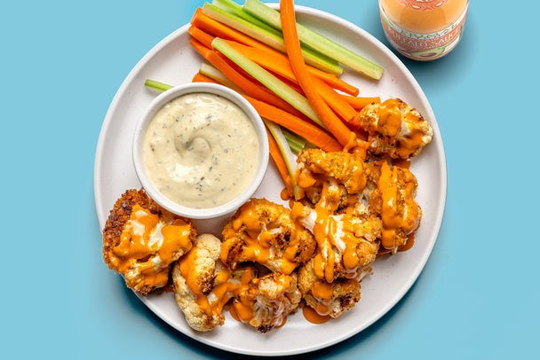 A plate of buffalo cauliflower wings drizzled in buffalo sauce with a side of ranch, on a blue table. 