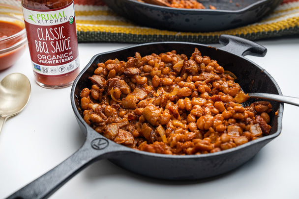 Baked beans with bacon and onion in a cast iron skillet, with a bottle of Primal Kitchen BBQ Sauce in the background. 