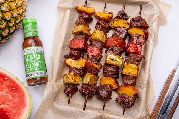 A tray of Hawaiian steak and fruit kabobs next to a bottle of Primal Kitchen Hawaiian Style BBQ Sauce and Marinade