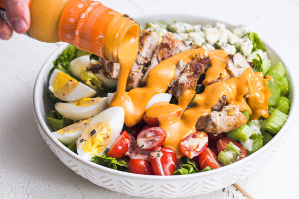 A bottle of Primal Kitchen Buffalo Sauce pouring onto a Chicken Cobb Salad in a large white bowl.