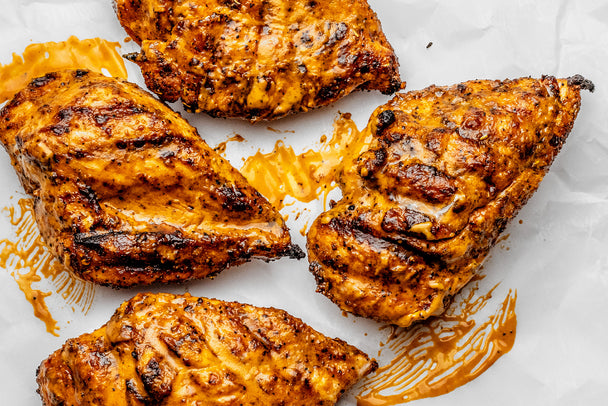 Grilled Buffalo Chicken Breast is on a white plate with a bowl of buffalo sauce, a brush, and a buffalo sauce bottle nearby.