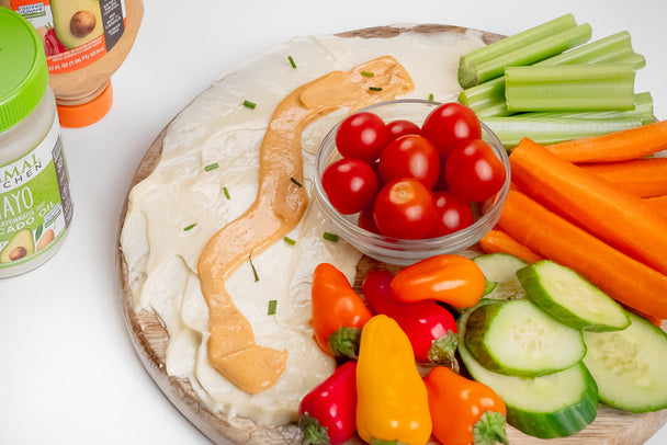 Primal Kitchen Avocado Oil Mayo and a swirl of Buffalo Mayo beside raw vegetables on a round wooden board.