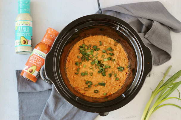 Buffalo Chicken Dip in a slow cooker. Bottles of Primal Kitchen Ranch and Buffalo Sauce in the background.