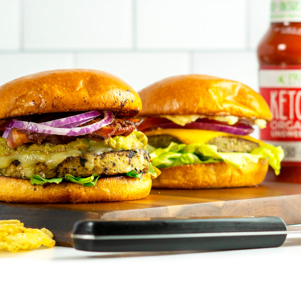 Two burgers are laid out on a bar with ketchup behind them.