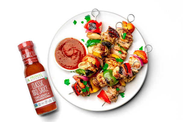 Grilled chicken and vegetable kabobs on a white plate next to a bottle of Primal Kitchen Classic BBQ Sauce