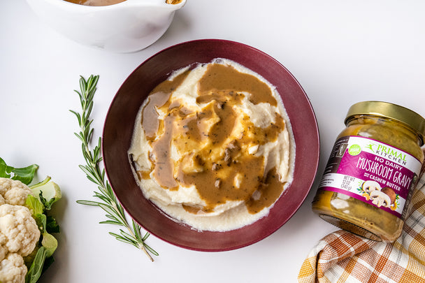 A brown bowl is filled with cauliflower mash topped with vegan mushroom gravy next to a jar of gravy and a sprig of rosemary.