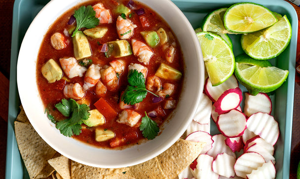 Mexican Shrimp Ceviche is in a white bowl on a blue serving tray with radishes, lime, chips, and with cocktail sauce and lime nearby.