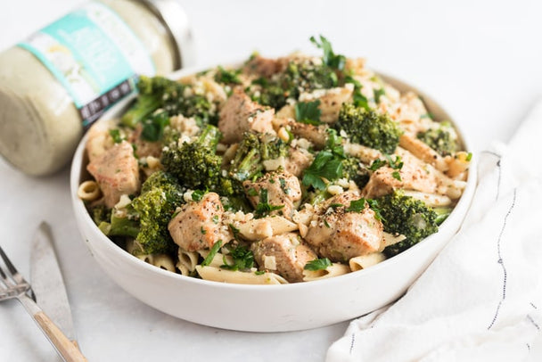 Broccoli florets, chicken, and penne pasta in Primal Kitchen Alfredo Sauce in a white ceramic bowl, next to a jar of sauce.