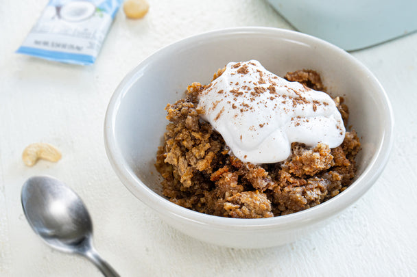 A white bowl filled with Baked Noatmeal with mocha latte flavor. Coconut whipped cream is on top of the baked noatmeal. A spoon is to the left of the bowl and a packet of Primal Kitchen Collagen Fuel Vanilla is on the side.