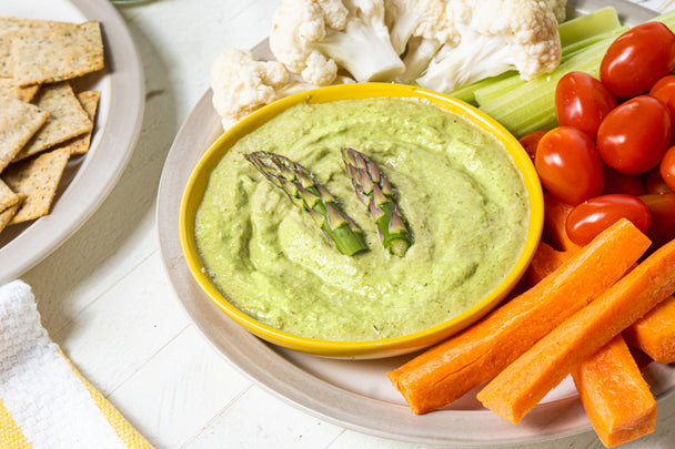 Asparagus dip in a beige bowl on a white plate with carrot sticks, cauliflower florets, celery sticks, and cherry tomatoes.