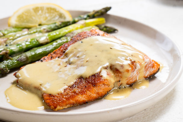 Salmon is drizzled in Hollandaise Sauce with asparagus and lemon on a white plate with a hollandaise sauce jar, yellow tea towel, and fork next to it.