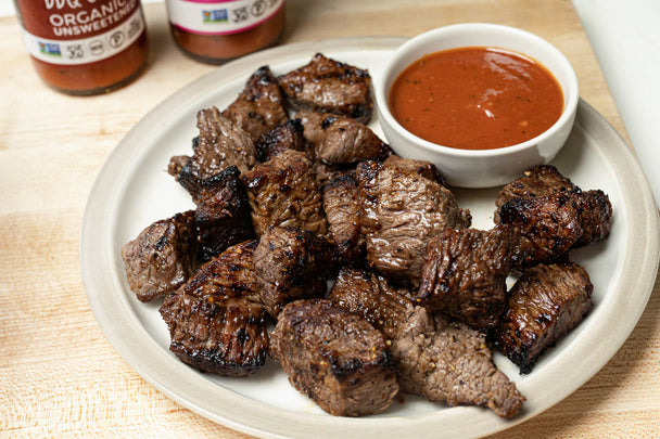 A white plate holds a serving of air fryer steak bites served with primal kitchen classic bbq sauce.