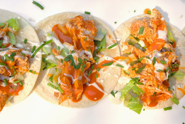 Three buffalo chicken tacos drizzled with Primal Kitchen Original Buffalo Sauce on a white plate. 