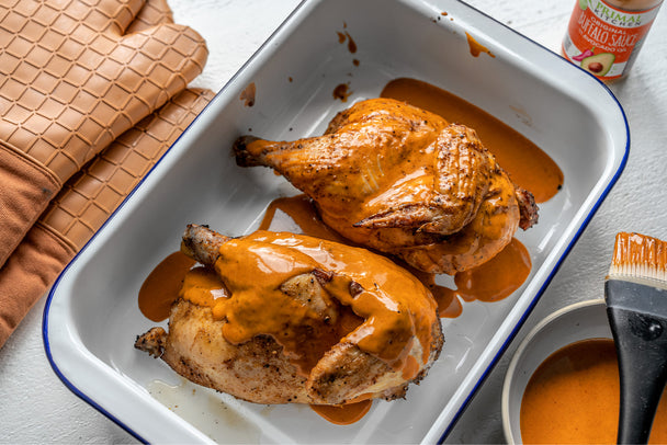 A pan of roasted chicken halves drizzled with Primal Kitchen Buffalo Sauce, oven mitts and sauce on the side