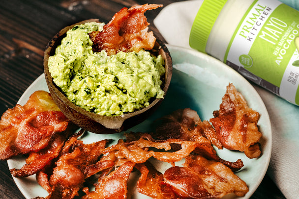 A plate of crispy bacon chips and a bowl of guacamole, with a jar of Primal Kitchen Mayo in the background.