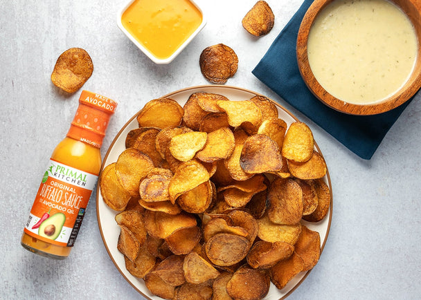 Homemade potato chips with Avocado Oil on a white plate, next to a bottle of Primal Kitchen Buffalo Sauce. 