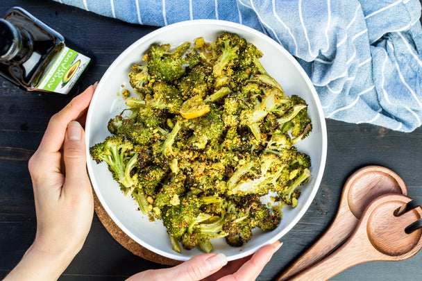 Hands cradling a big white bowl filled with roasted broccoli with cheese sauce. Next to the bowl are wooden spoons, Primal Kitchen Avocado Oil, and a blue napkin with white stripes. 