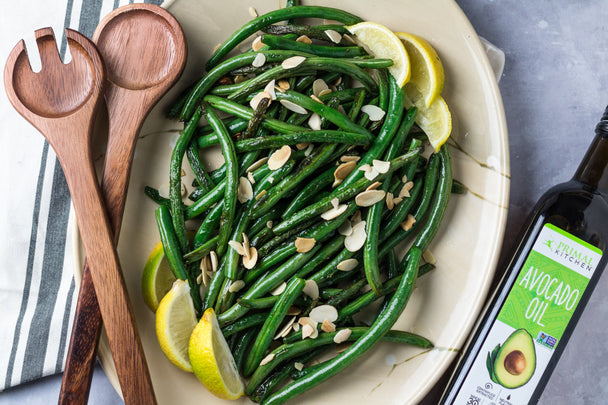 Sauteed green beans with sliced almonds and lemon wedges on a large white platter. Brown wooden spoons are resting on the plate. Primal Kitchen Avocado Oil is next to the plate. 