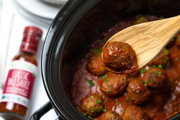 BBQ Meatball on a wooden spoon held over a slow cooker filled with meatballs coated in Primal Kitchen Classic BBQ Sauce and garnished with chopped parsley