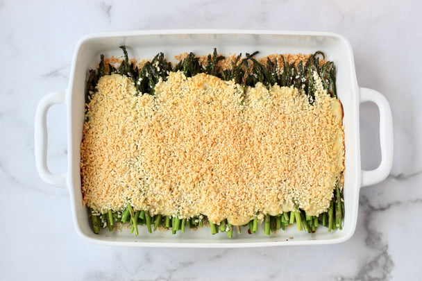 A white ceramic dish of cooked asparagus casserole topped with cheese and breadcrumbs, on a marbled background.