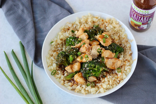 A bowl of rice topped with Teriyaki Chicken, with green onions, a linen napkin, and a bottle of Thick & Sticky Honey Teriyaki Sauce in the background.