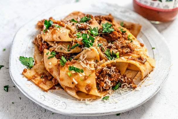 Pappardelle with spicy arrabbiata meat sauce on a white plate next to tomatoes and Primal Kitchen Arrabbiata Sauce