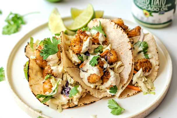 Three shrimp tacos on a white plate with lime wedges and Primal Kitchen Pesto Mayo in the background