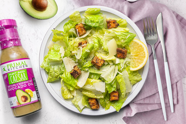 A plate of plant based caesar salad next to a bottle of Primal Kitchen Plant Based Caesar Dressing