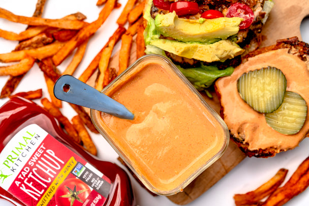 An open-faced burger with a container of secret sauce next to a squeeze bottle of Primal Kitchen A Tad Sweet Ketchup.