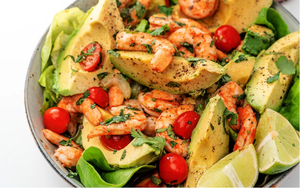 A close-up of shrimp, lime wedges, avocado wedges, and cherry tomatoes seasoned with herbs in a white bowl.