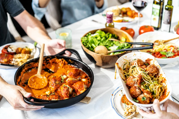 Many hands are placing dishes on a table full of food.