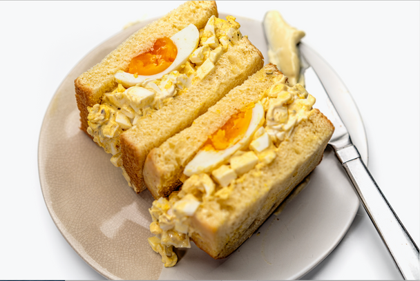 An egg salad sandwich is on a cream colored plate with a knife laying nearby.