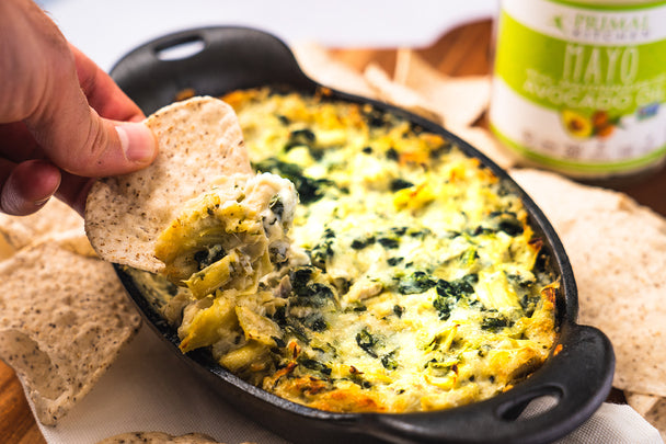 A man's hand dipping a tortilla chip into baked spinach artichoke dip in a cast iron dish. Tortilla chips and Primal Kitchen Mayo in the background.