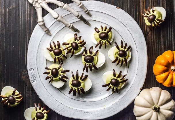 Spooky Deviled Eggs on a white plate with more eggs and little pumpkins in the background. 