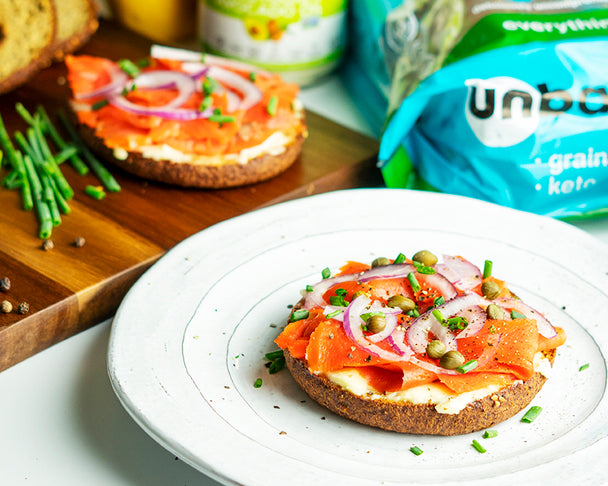 A smoked Salmon breakfast bagel is prepared on a white plate with a cutting board with ingredients, including unbun bagels, is in the background.