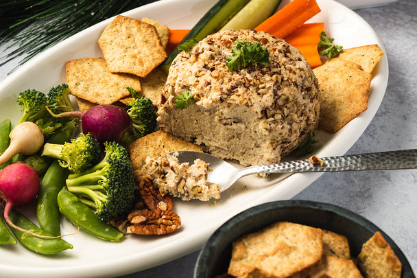 Close up shot of vegan cheese ball with a silver spoon scooping out the cheese. Also on the white plate: broccoli florets, radishes, snap peas, carrot and cucumber sticks, and crackers.