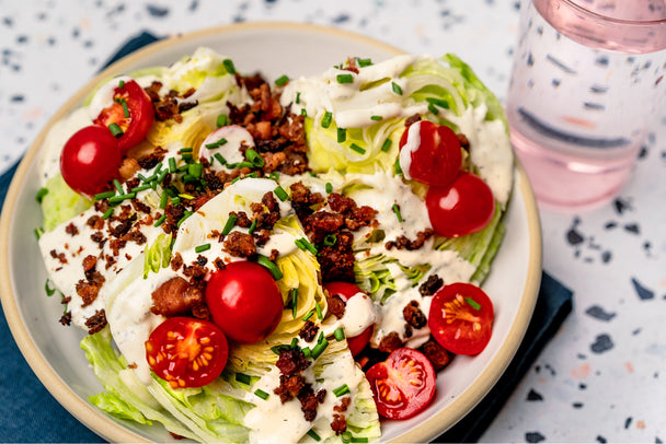 A BLT Wedge Salad drizzled with Primal Kitchen Ranch Dressing on a beige plate, with a pink glass and a speckled countertop in the background.