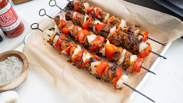 Four kabobs with onion, red pepper and steak are laid out on a metal pan covered with parchment, while a wooden bowl of salt and a ketchup bottle are nearby.