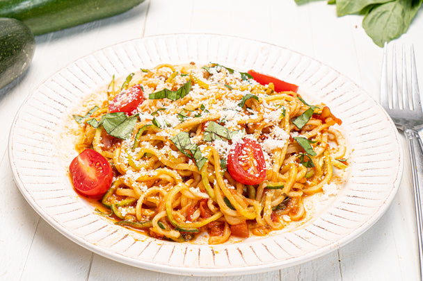 Zoodles with Primal Kitchen marinara sauce, sliced cherry tomatoes, chopped basil, and parmesan cheese on a white plate