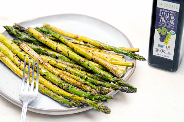 Balsamic glazed asparagus on a white plate with fork next to Primal Kitchen Balsamic Vinegar of Modena bottle