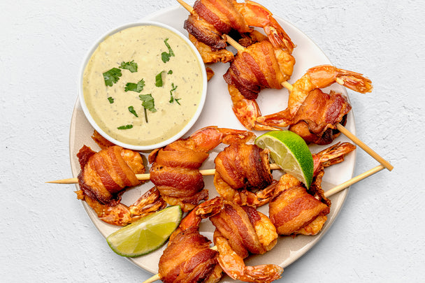 Three skewers of bacon-wrapped shrimp with lime wedges and a dipping bowl of Primal Kitchen Avocado Lime Ranch Dressing, on a white plate. 