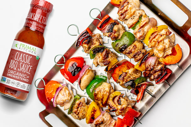 5 Grilled Chicken kabobs with vegetables are laid out on a metal tray with a bottle of BBQ Sauce next to it.