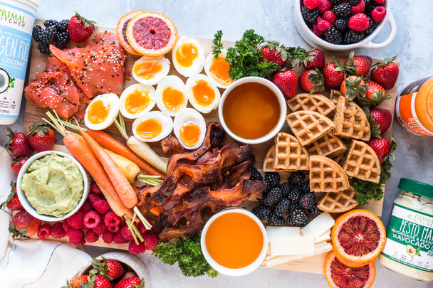 A breakfast charcuterie board is assembled on a marble countertop with mayo, collagen, and buffalo sauce nearby.