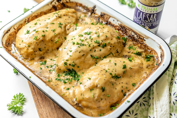 Baked Caesar chicken breasts in a white casserole dish with Primal Kitchen Caesar Dressing on the side.