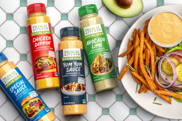 Four Primal Kitchen Dipping Sauces on a tiled background next to a burger and fries on a plate.