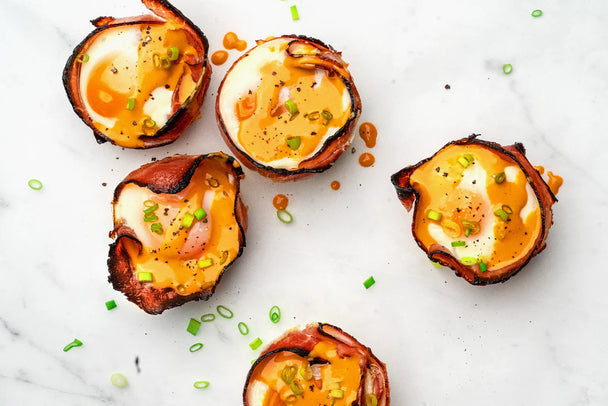 5 ham and egg cups sprinkled with chopped green onion and topped with Primal Kitchen Buffalo Sauce on a round white plate. A jar of Primal Kitchen Buffalo Sauce is next to the plate.