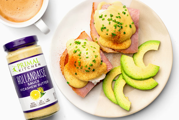 Keto eggs benedict: sliced ham with poached eggs, hollandaise sauce, chopped chives, and paprika on a white plate. Avocado slices are also on the plate. Next to the plate is a jar of Primal Kitchen Hollandaise Sauce.