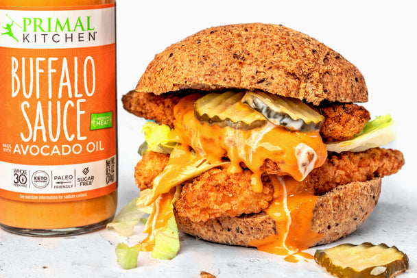 Spicy chicken sandwich with a keto bun, keto chicken tenders, iceberg lettuce, pickles, and Primal Kitchen Buffalo Sauce. Sandwich next to a bottle of Primal Kitchen Buffalo Sauce.